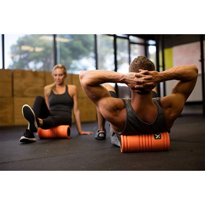 "A man and woman in a gym uses the TriggerPoint GRID® Foam Roller (orange) on her leg to release muscle pain and tightness." 